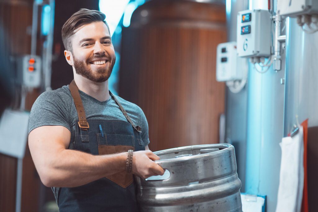 A young brewer in an apron holds a barrel with beer in the hands of a brewery.